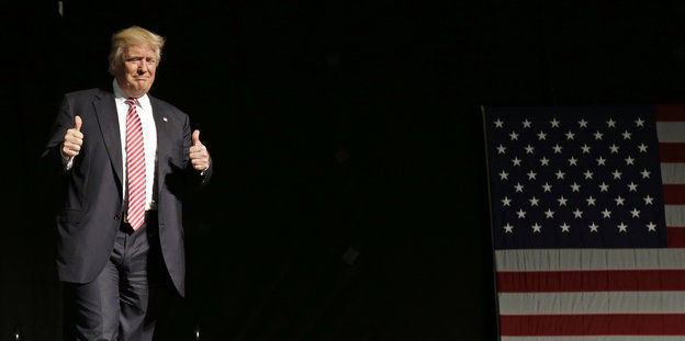Donald Trump steht auf der Bühne vor einer USA-Flagge, verzieht das Gesicht und hält beide Daumen hoch