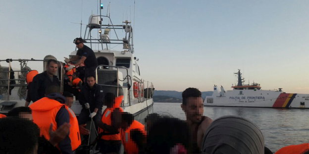 Flüchtlinge mit Mitgliedern der Küstenwache in orangefarbenen Warnwesten auf einem Boot, im Hintergrund das Frontex-Boot