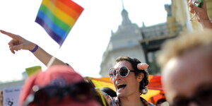 Mann mit Sonnenbrille, Penishaarreif und Discokugel-Ohrringen auf einer Gay-Pride in Sofia, Bulgarien. Im Hintergrund ist eine Regenbogenflagge.