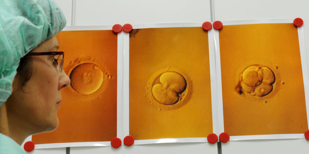 Eine Frau in Laborkleidung vor der Abbildung der Teilung einer befruchteten Eizelle
