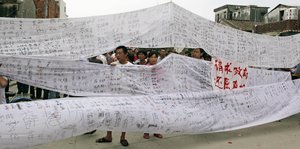 Menschen tragen weiße Stoffbahnen, auf denen chinesische Schriftzeichen zu sehen sind