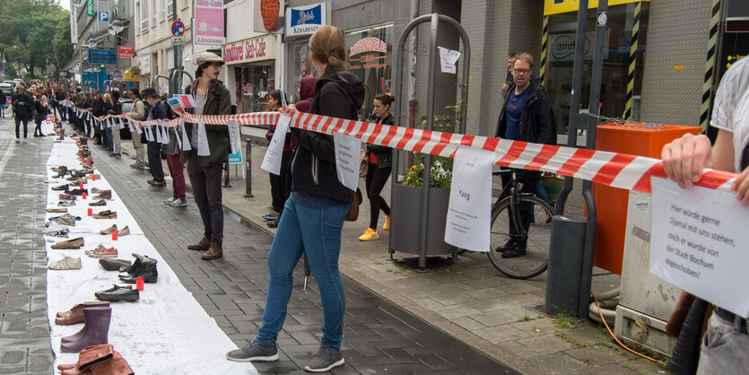 Menschen stehen entlang einem rot-weißen Absperrband, davor eine weißer Streifen Papier, auf dem Schuhe aufgereiht sind