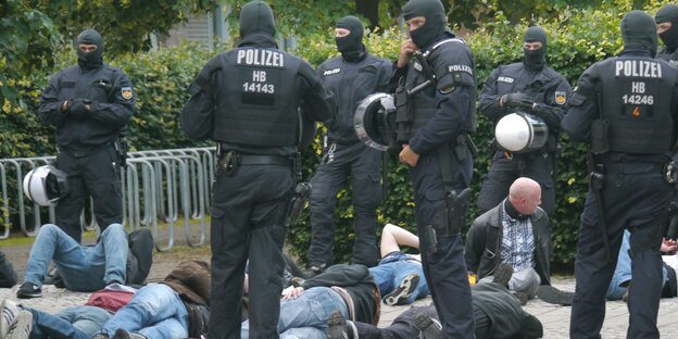 Vermummte Polizisten stehen neben Personen, die auf dem Boden liegen