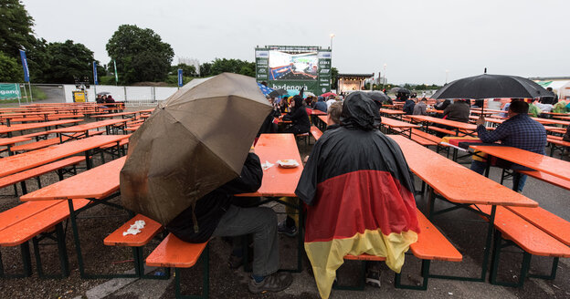 Zwei deutsche Fans sitzen auf Bierbänken. Einer mit Regenschirm, der andere mit einem Regencape