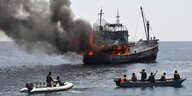 Ein Fischerboot auf dem offenen Meer in Flammen, davor zwei kleinere Boote