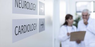 Zwie Ärzte unscharf im Hintergrund, im Vordergrund ein Schild, das zur Kardiologie weist