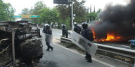 Polizisten in Kampfmontur auf einer Straße, auf der ein Auto brennt