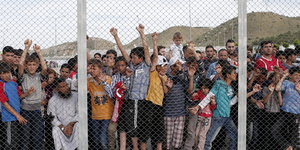 Menschen, darunter viele Kinder, hinter einem Zaun, im Hintergrund Hügel