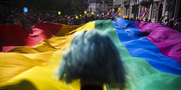 Eine Person mit türkisfarbenen Haar steht vor einer großen Regenbogenfahne