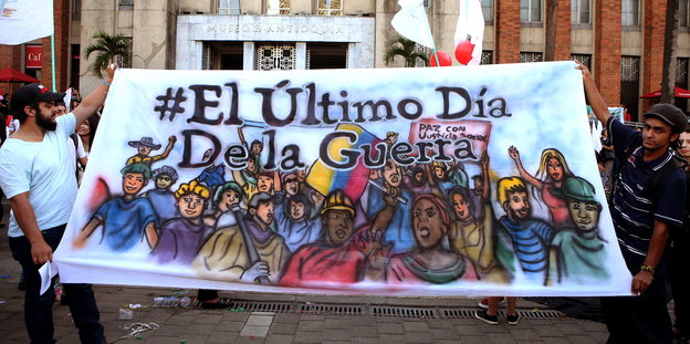 zwei Männer halten ein buntes Poster mit der Aufschrift „#El Último Día de La Guerra“