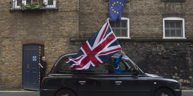 Aus einem britischen schwarzen Taxi hält jemand eine große britische Flagge. Über dem Auto hängt aus einem Fenster eine EU-Flagge