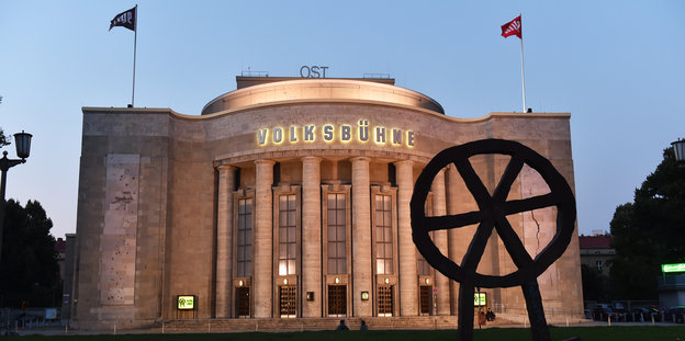 Das Gebäude der Berliner Volksbühne mit sechs Säulen in der Abenddämmerung. Im Vordergund das Profil eines Rads mit Speichen