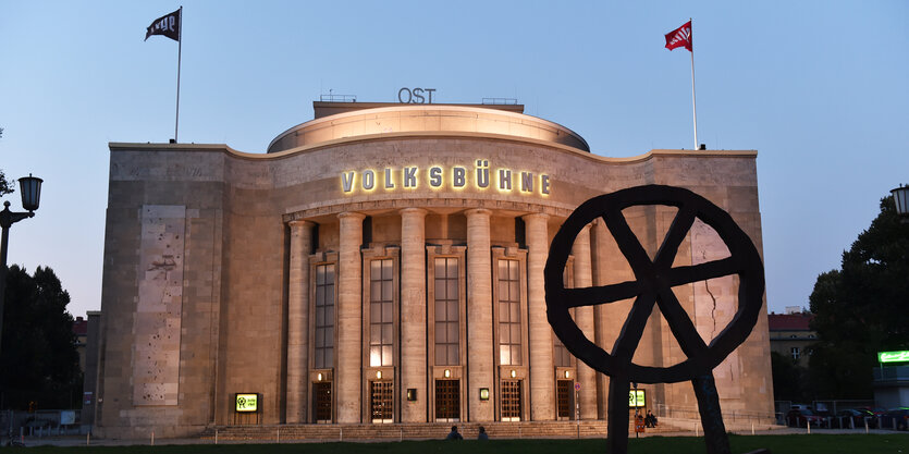 Das Gebäude der Berliner Volksbühne mit sechs Säulen in der Abenddämmerung. Im Vordergund das Profil eines Rads mit Speichen