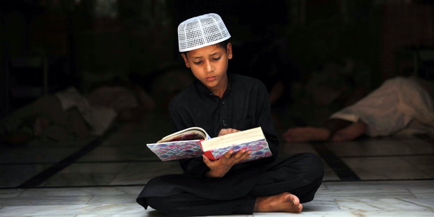 Ein Junge in dunklem Gewand und weißer Mütze ließt, mit überkreuzten Beinen sitzend, den Koran