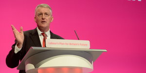 Hilary Benn vor rosafarbenem Hintergrund an einem Rednerpult