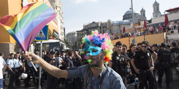 Ein Mann trägt eine Maske und Perücke und schwingt ein Fahne in Regenbogenfarben, im Hintergrund Polizisten