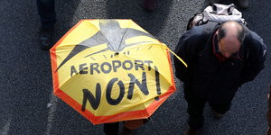 Ein Demonstrant trägt einen Schirm, auf dem eine Parole gegen den Flughafen steht