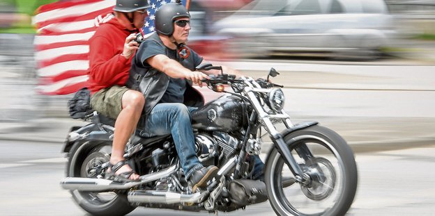 Zwei Männer fahren auf einer Harley Davidson mit einer US-Flagge.