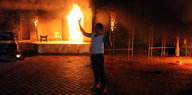 Ein Mann hält nachts ein Gewehr in die Luft, hinter im brennt ein Gebäude