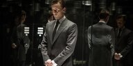 Ein Mann in grauem Anzug steht mit verschränkten Händen in einem verspiegelten Fahrstuhl