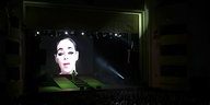 Ein Frauengesicht auf einer großen Leinwand, vor ihr auf der Bühne eine grüne Gestalt