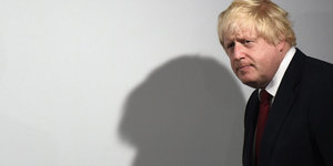 Ein Mann guckt nachdenklich. An der Wand ist sein Schatten abgebildet. Es ist Boris Johnson.