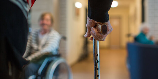 Zwei ältere Menschen auf einem Flur: einer sitzt im Rollstuhl, der andere hat einen Gehstock in der Hand