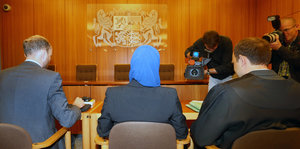Zwei Männer und eine Frau mit Kopftuch in einem Gerichtssaal