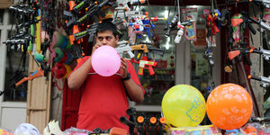 Ein Man bläst an einem Verkaufsstand Luftballons auf, hinter ihm hängen Spielzeugwaffen