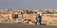 Zwei Mädchen laufen mit einem Esel eine Straße vor einer Siedlung entland
