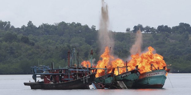 Ein Boot liegt in Flammend stehend auf dem Wasser, dahinter ein Wald