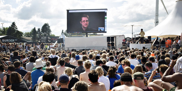 Eine Menschenmenge unter freiem Himmel schaut auf eine Videoleinwand, auf der Snowden zu sehen ist