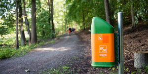Neben einem Waldweg hängt ein grün-orangefarbener Mülleimer