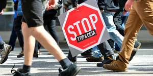 Menschen laufen über eine Straße. Eine Person hält ein Schild auf dem Stop TTIP und Ceta steht