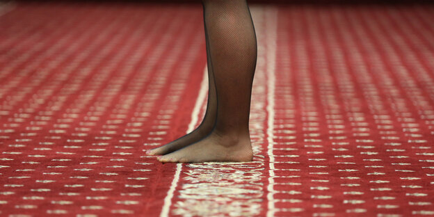 Auf einem rot-gemusterten Teppich stehen Beine in schwarzen Strumpfhosen