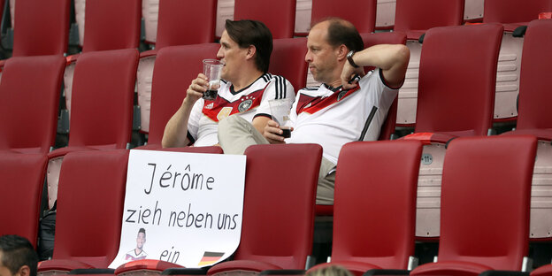 Zwei Deutschland-Fans sitzen im Stadion vor einem Transparent, auf dem steht: "Jerome, zieh neben uns ein."
