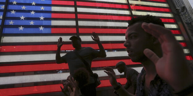 Vor dem Hintergrund einer US-Flagge gehen Menschen mit erhobenen Händen