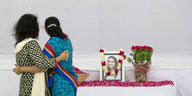 Zwei Frauen halten sich in den Armen und blicken auf ein Trauerfoto neben einem Blumenstrauß