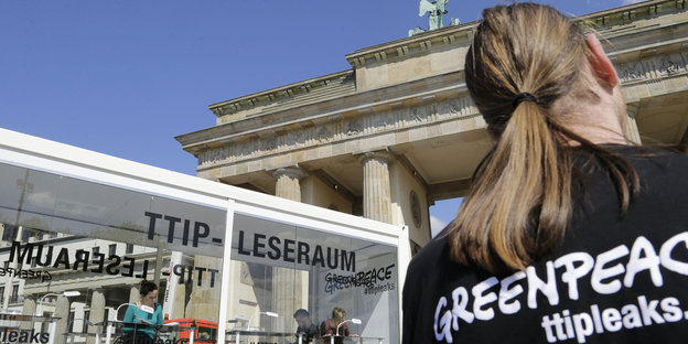 Szene vor dem Brandenburger Tor: Ein Glaskasten und ein Mensch von hinten mit langem Zopf, auf dem Tshirt die Aufschrift Greenpeace ttipleaks