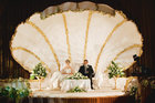 In einer beleuchteten weißen Muschel sitzt ein Hochzeitspaar, eine weiße Treppe führt zu ihnen hinauf, der Raum ist dunkel