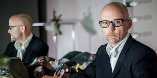 Moby sitzt im Anzug vor einem Spiegel, neben ihm ist sein Spiegelbild zu sehen