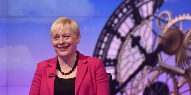 Angela Eagle in einem Fernsehstudio vor einer dekorativen Uhr