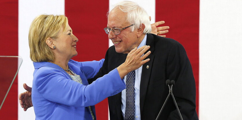 Umarmen sich auf einer Bühne vor der US-Fahne: Bernie Sanders und Hillary Clinton