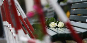 Zwei Weiße Rosen liegen auf einer Bank, davor sieht man ein rot-weißes Absperrgitter