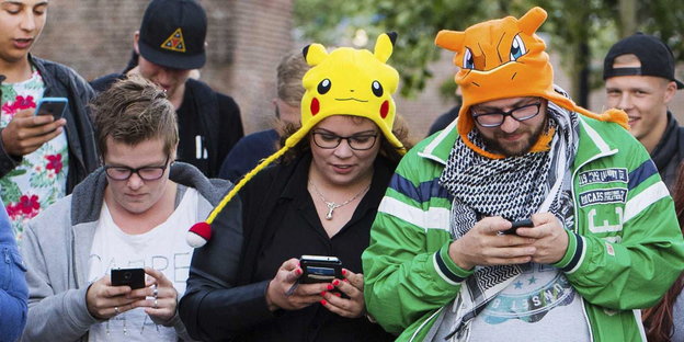 Mehrere junge Leute starren auf ihr Smartphone, zwei tragen Mützen in Pokémon-Optik