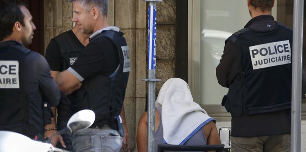 Französische Polizisten stehen um einen Mann, der ein Handtuch auf dem Kopf trägt
