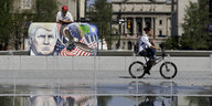 Auf einer Leinwand ist ein gesprühtes Kunstwerk mit dem Gesicht Donald Trumps und einer Weltkugel zu sehen, im Vordergrund fährt ein Radfahrer an einem Springbrunnen vorbei