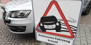 Auf einem Schild steht „Autonomes Auto kreuzt“. Dahinter steht ein Auto