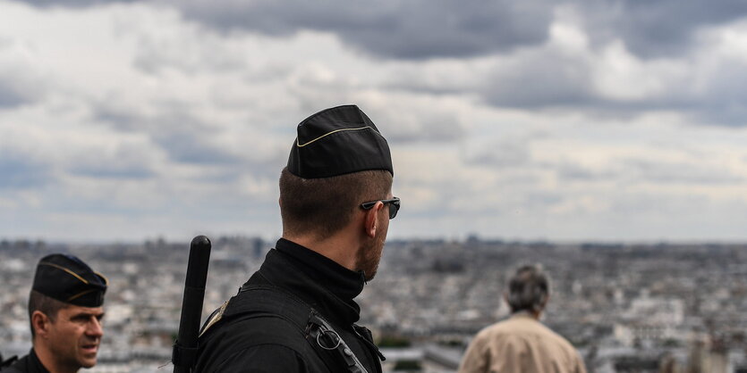 Polizisten mit Käppis, einer im Vorder-, der andere im Hintergrund. In der Ferne die Stadtkulisse von Paris