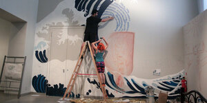 Eine Frau und ein Mann auf einer Leiter arbeiten an einem Wandbild, das eine Welle zeigt